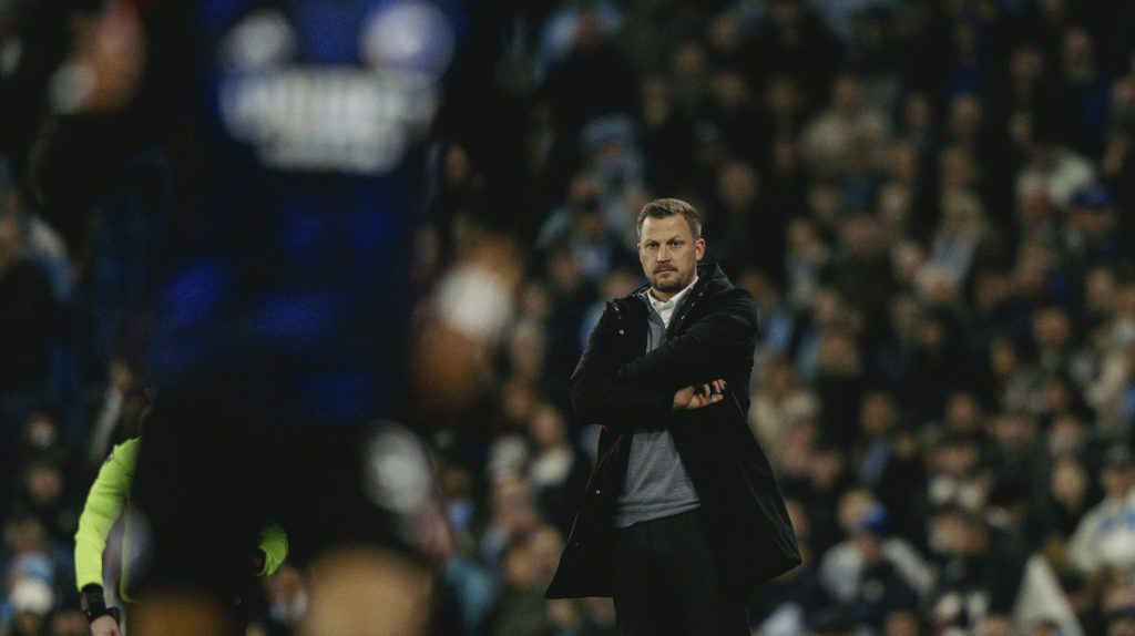 Jacob Neestrup står i sort jakke med armene over kors og ser ind på banen. Bag ham er masser af tilskuere, og i forgrunden af billede en meget utydelig FCK spiller.